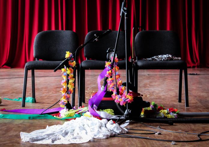 Three black folding chairs sitting in front of a red Theatre curtain, with various microphone stands decorated with flowers and other costume accessories.