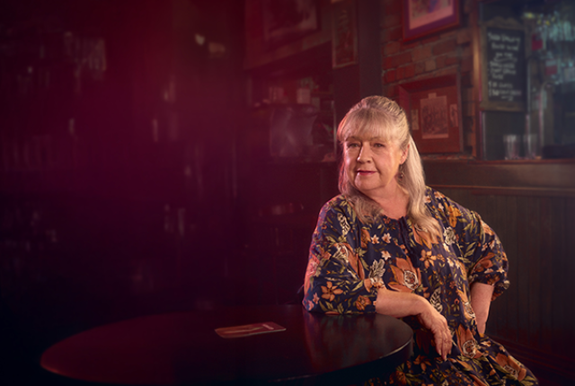 A woman leans on a table, with a bar in the background