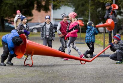 Children in winter coats playing with a giant red megaphone in an outdoor park on a footpath. One is speaking into one end of the megaphone, while the other listens at the other end. while
