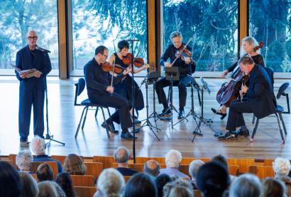 Five seated musicians on a stage performing various string instruments, with a man standing in front of a microphone holding a large book